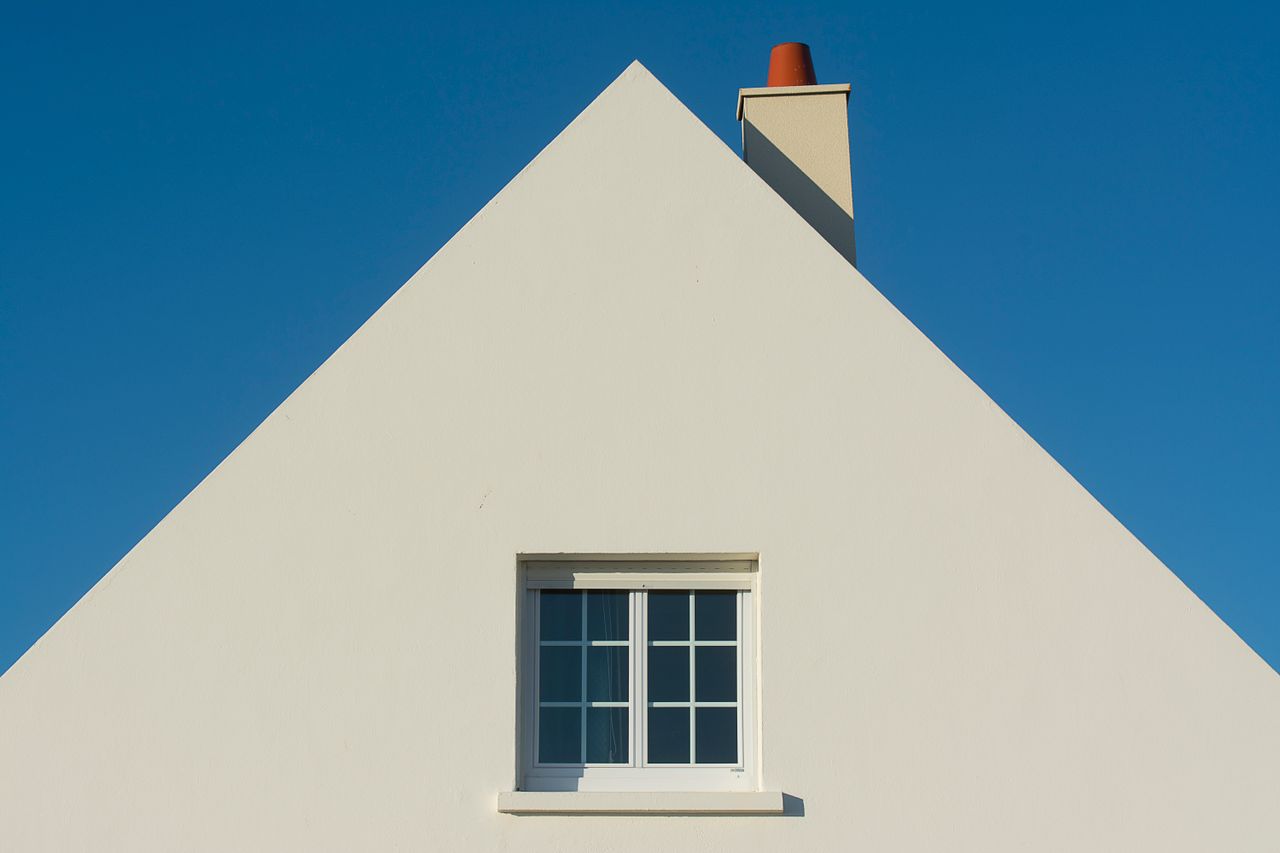 Simple triangular house facade with window and chimney against a clear blue sky.